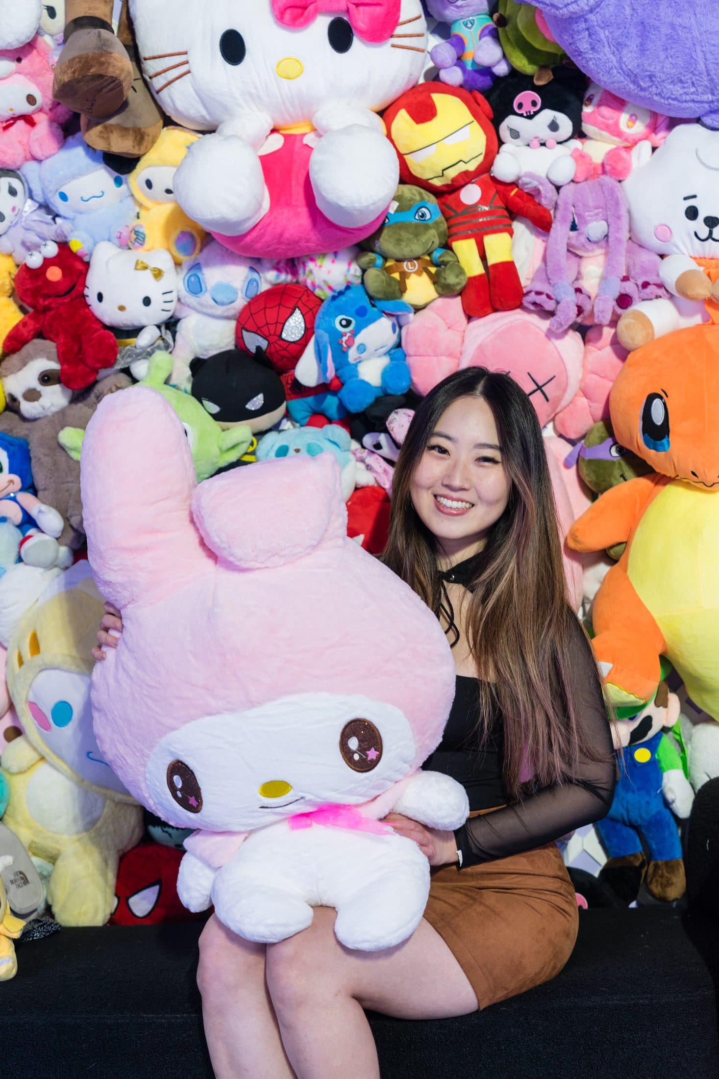 A guest sitting in front of a wall of colourful plush prizes, holding a large plush.