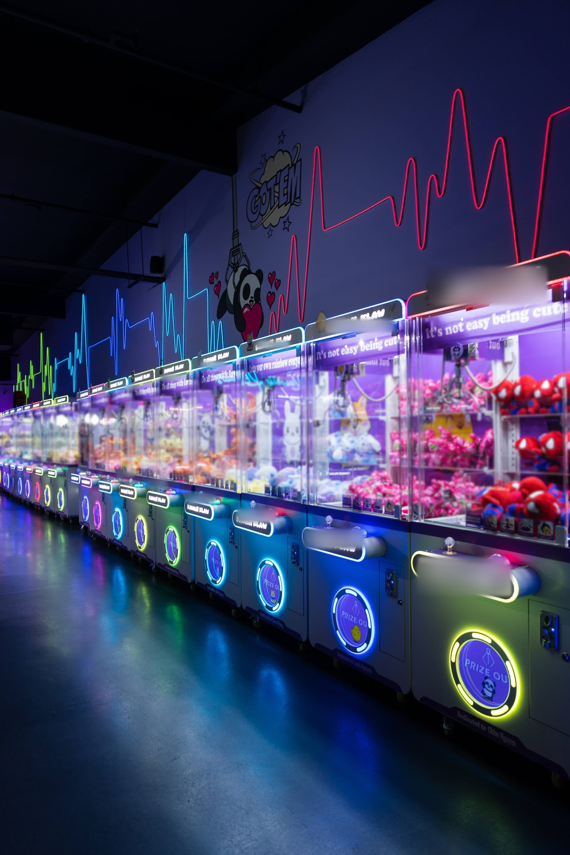 A long aisle of claw machines glowing pink, blue and green in a darkened arcade, reflected in the polished floor.