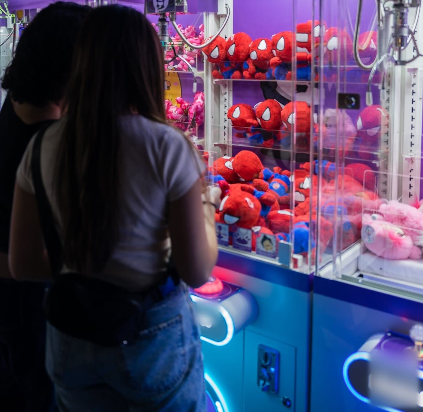A player at the controls of a brightly lit claw machine filled with red plush toys.